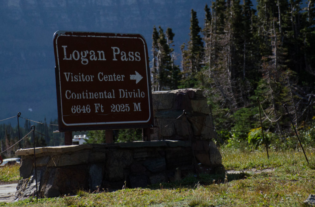 Sign for Logan Pass, Glacier National Park, shows direction of visitor center and 6646' height at Continental Divide